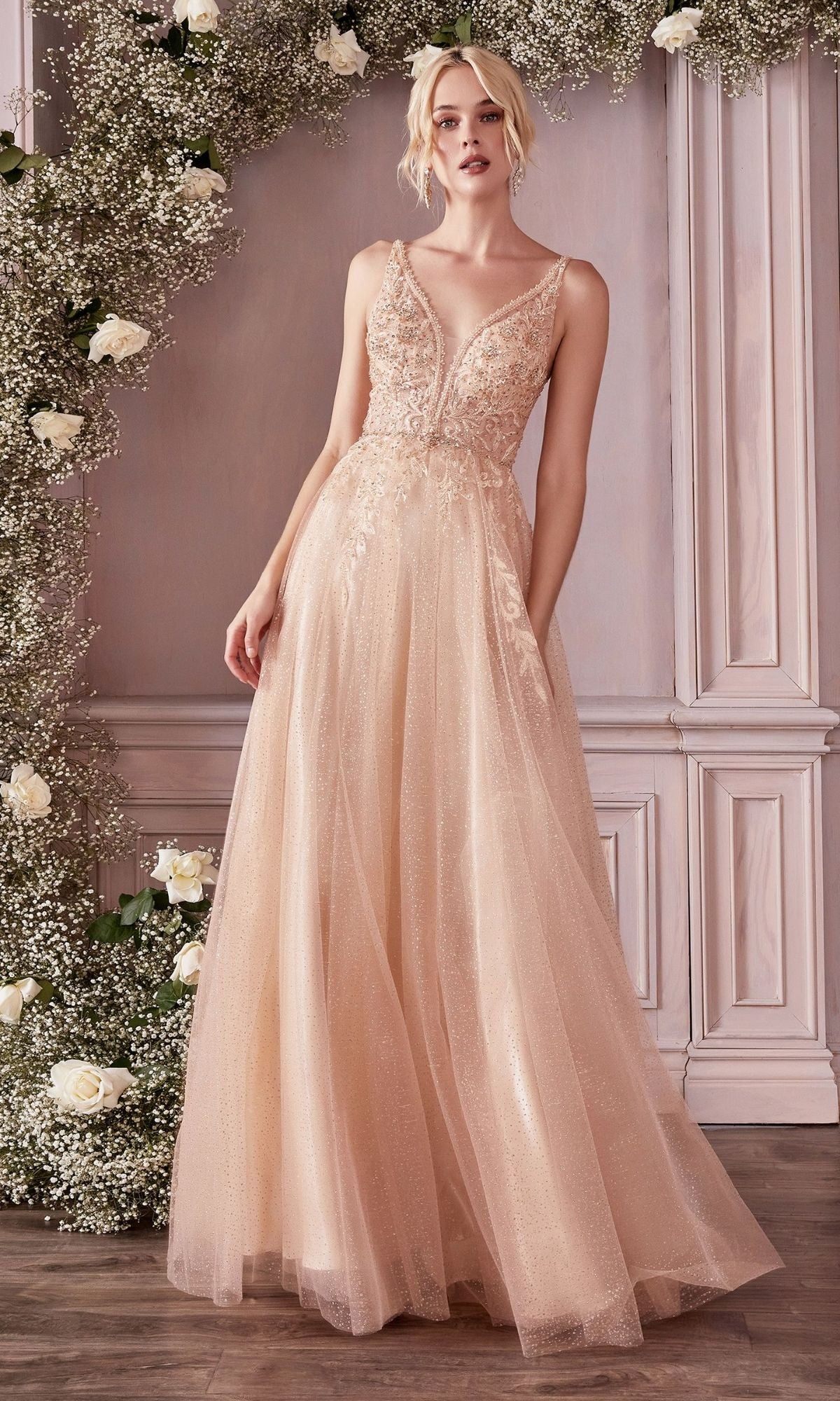 A woman in the Beaded-Bodice Long Pastel A-Line Prom Dress CD0196 stands indoors beside a floral arrangement of white roses and greenery. The dress features a blush pink hue, sleeveless design, V-neckline, and lace details.